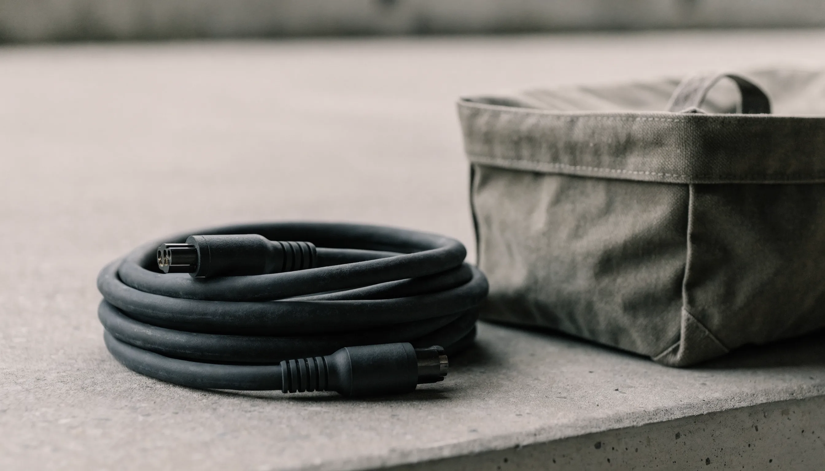 A neatly coiled heavy-duty charging cable beside a canvas storage bag on a plain concrete surface.
