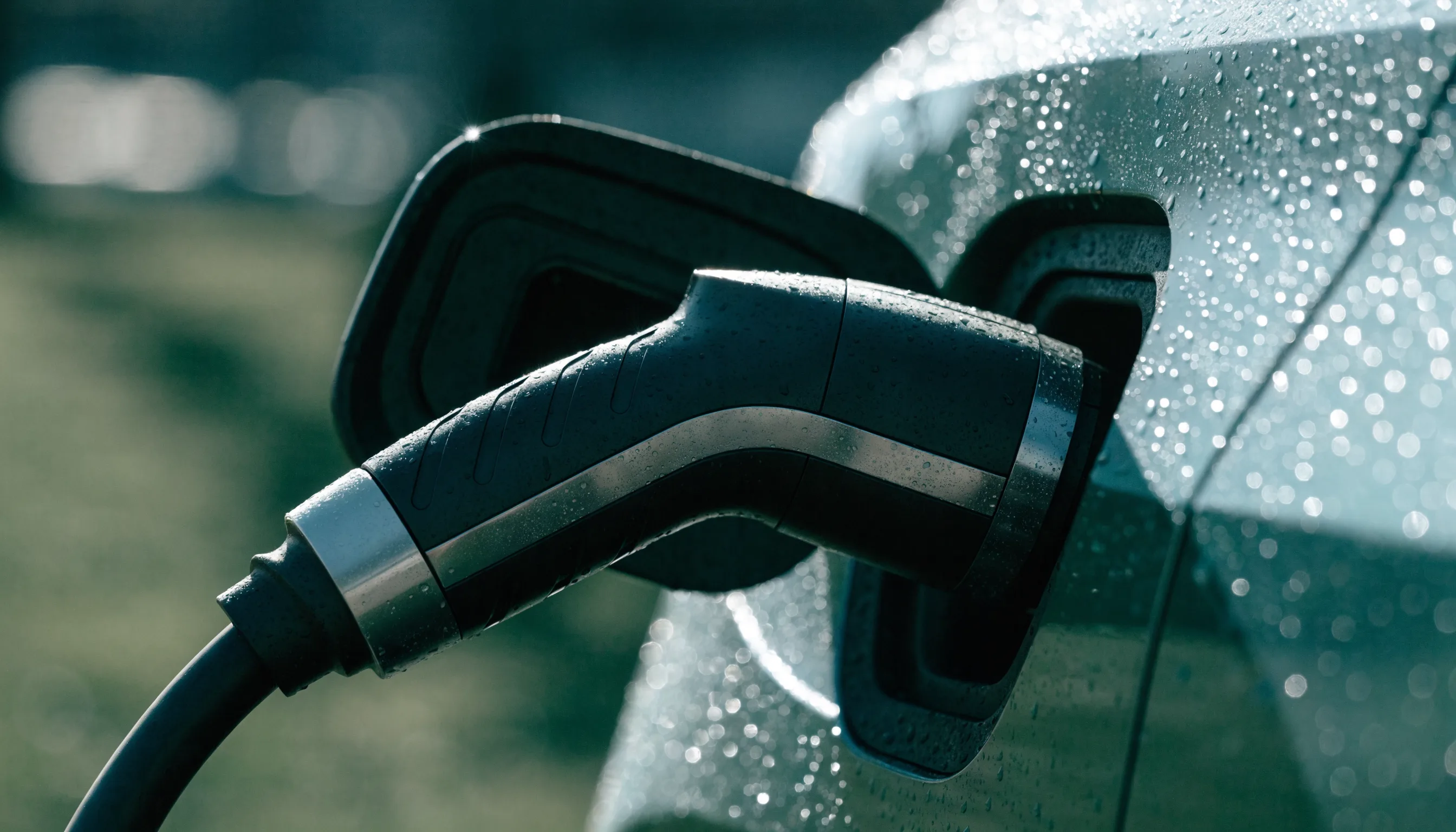 Close-up of an electric vehicle charging connector plugged into a car's charge port, with water droplets on the surrounding paintwork.