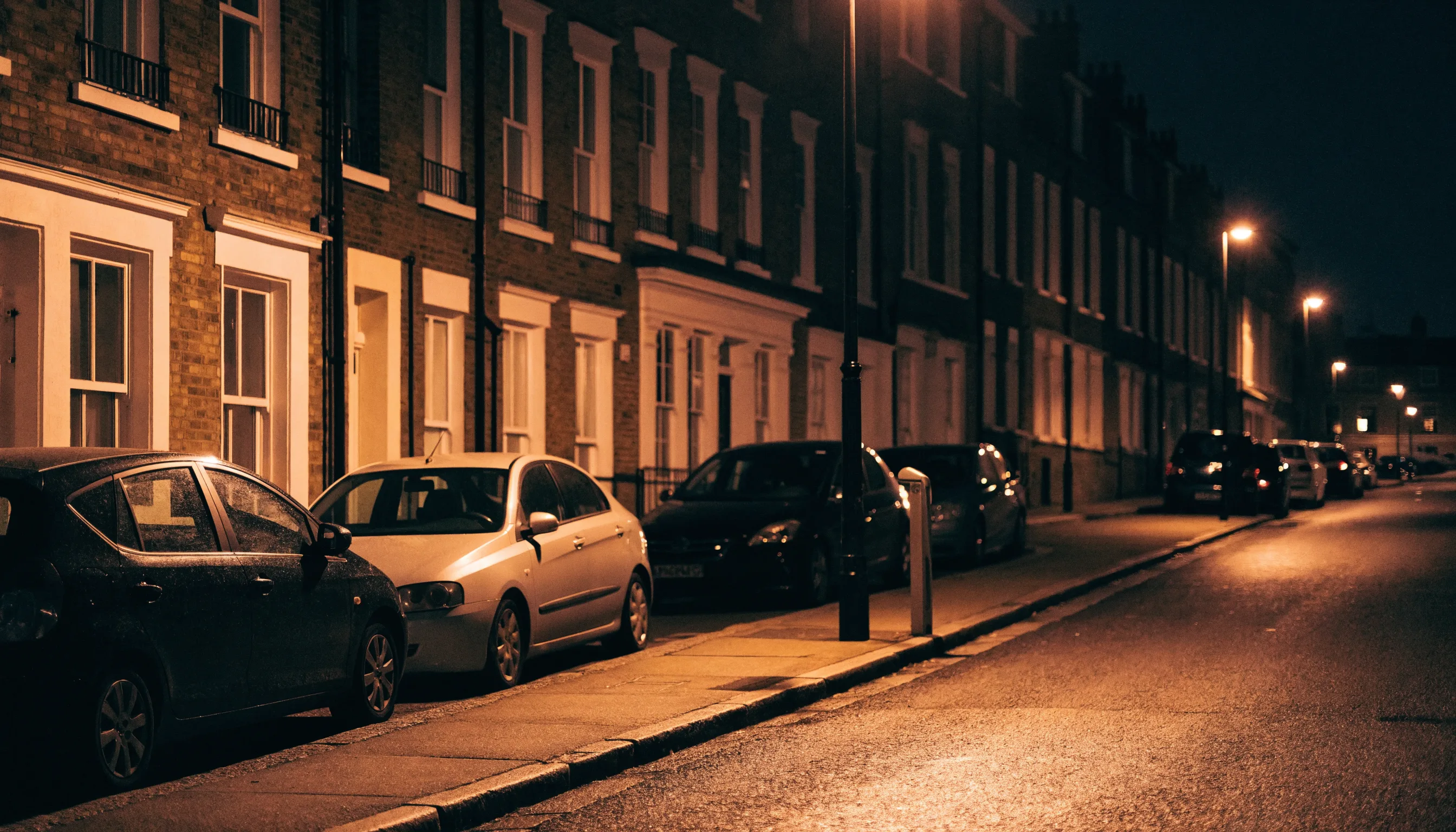 A terraced residential street at night with cars parked along the kerb and a slim charging bollard beside a lamp post.