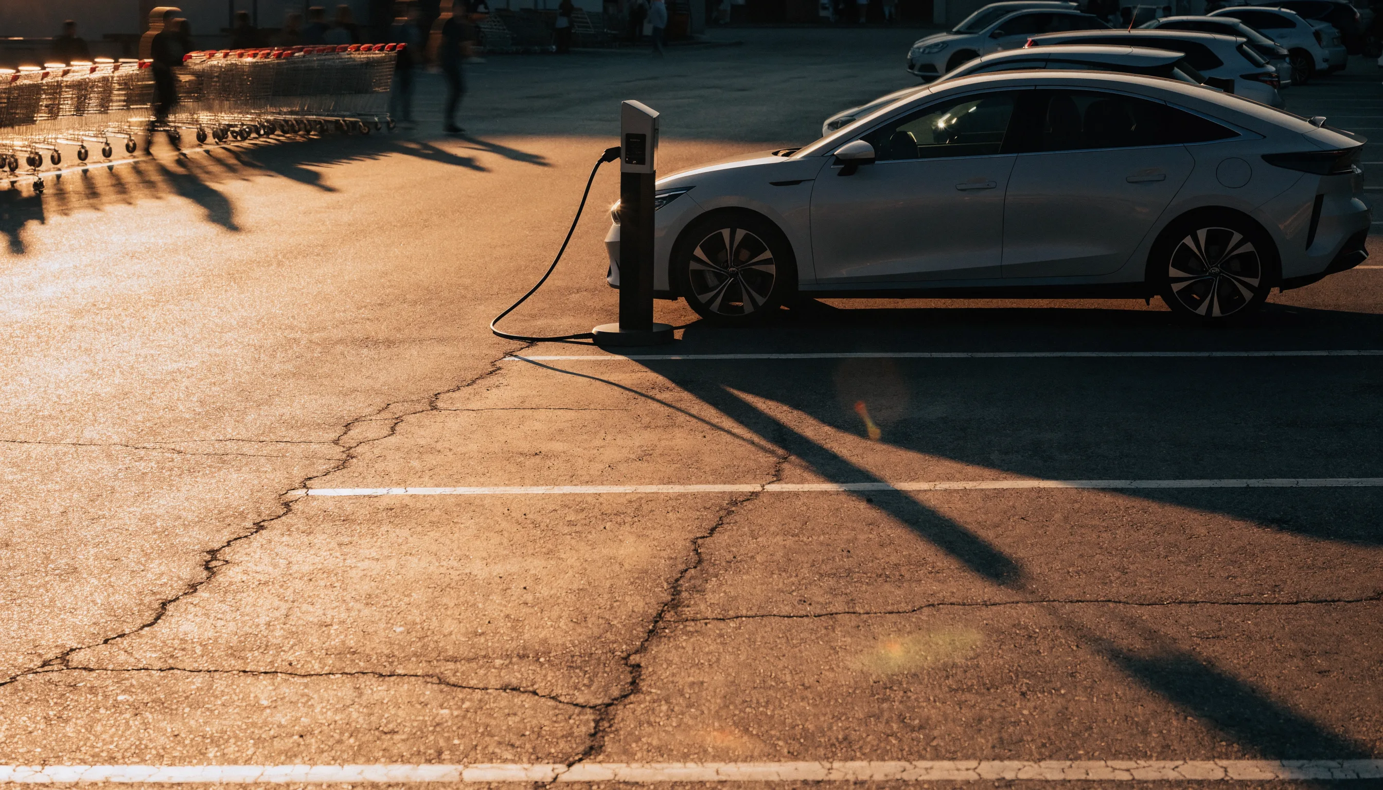 An electric family car plugged into a charging post in a supermarket car park during golden hour.