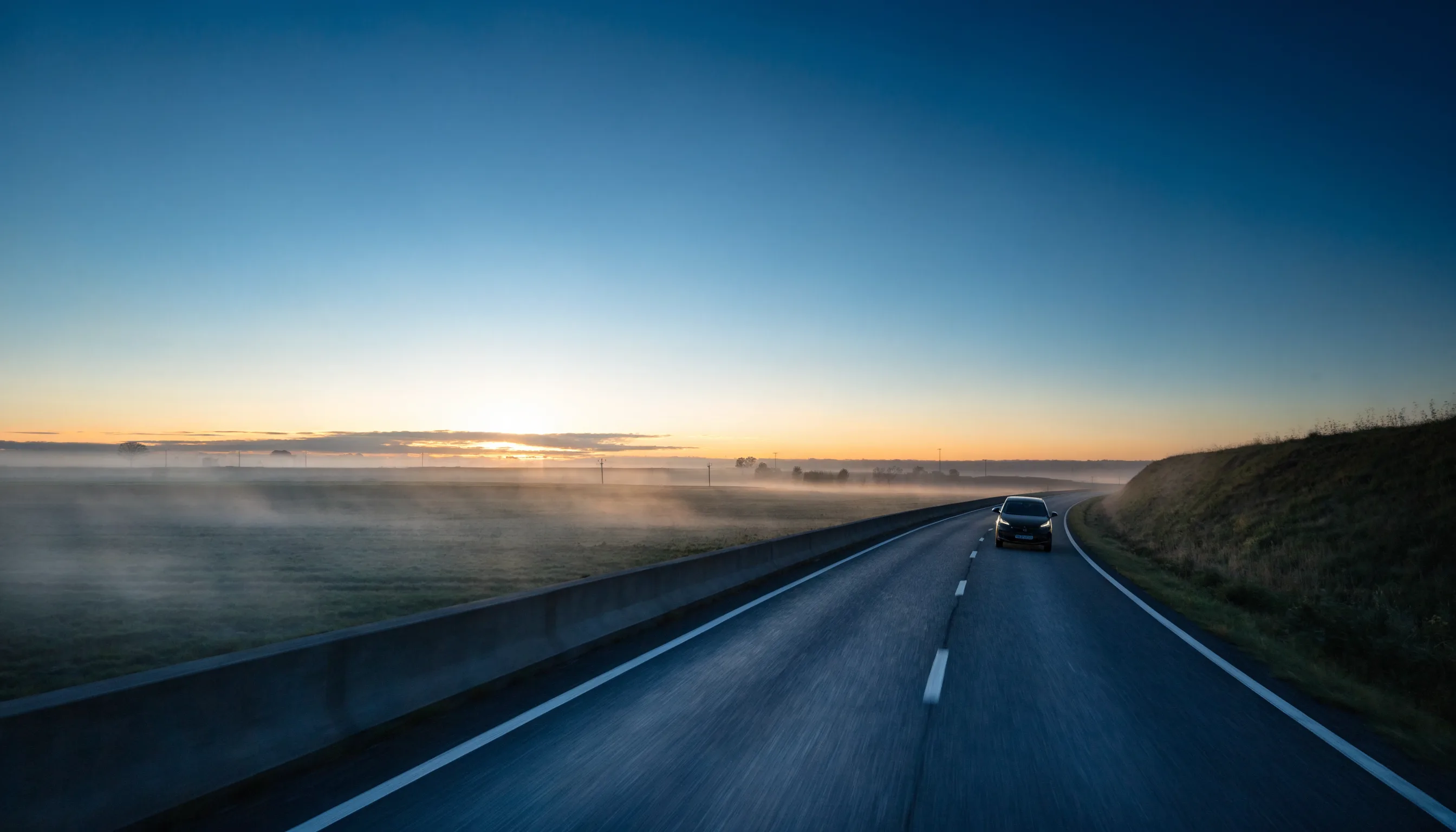 An electric car travelling along an open motorway at dawn, with mist across the surrounding fields.