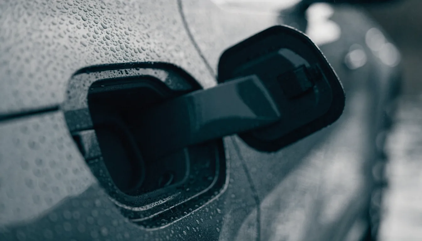 An open electric car charge port door in the rain, with water beading on the surrounding paintwork.