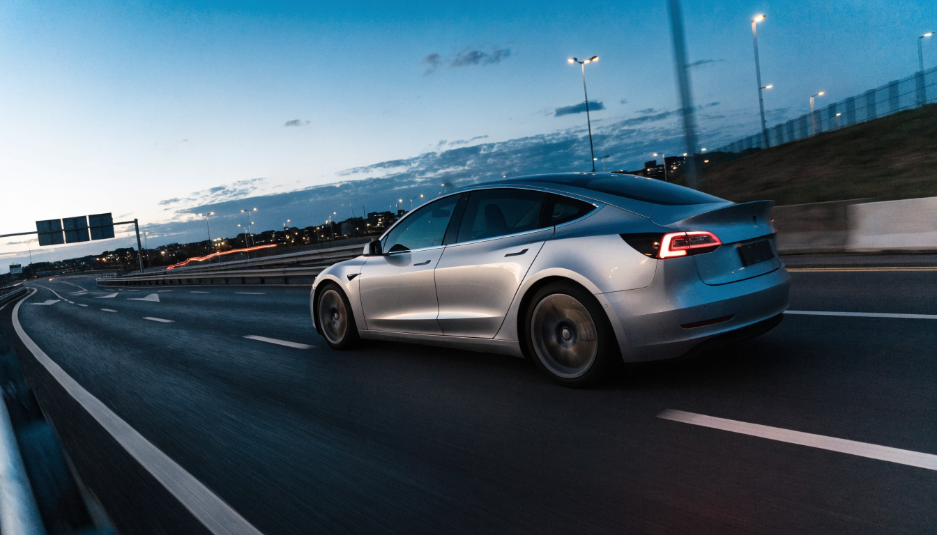 A silver Tesla Model 3 accelerating away from a junction on an empty UK dual carriageway at dusk, its wheels slightly blurred with motion.